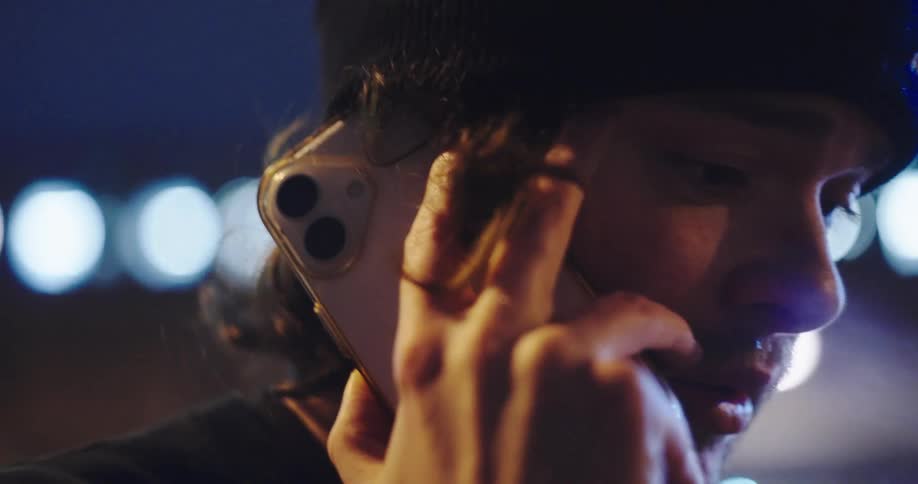 Moody close-up shot of a young man wearing a beanie talking on his smartphone at night