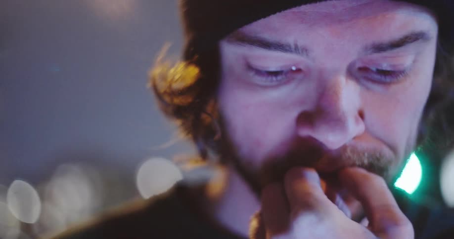 A dramatic slow-motion close-up reveals a serious young man in a beanie, lost in thought under the city's night lights