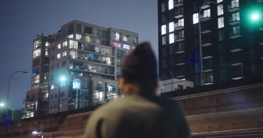 A person wearing a beanie stands on a city street at night, looking out at illuminated apartment buildings