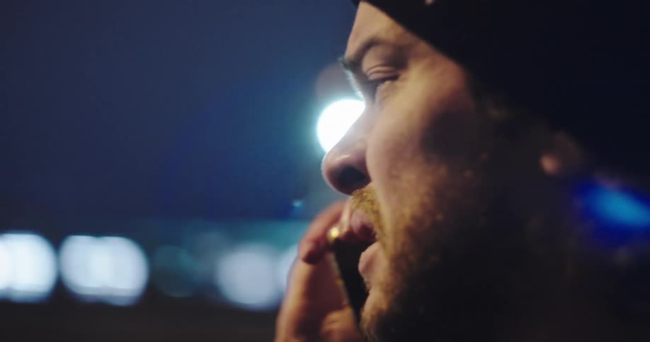 A pensive man wearing a beanie has a difficult conversation on his phone at night, illuminated by passing city lights