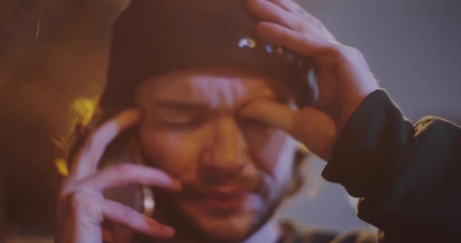 An extremely distressed man with a beanie has an emotional phone call on the street at night