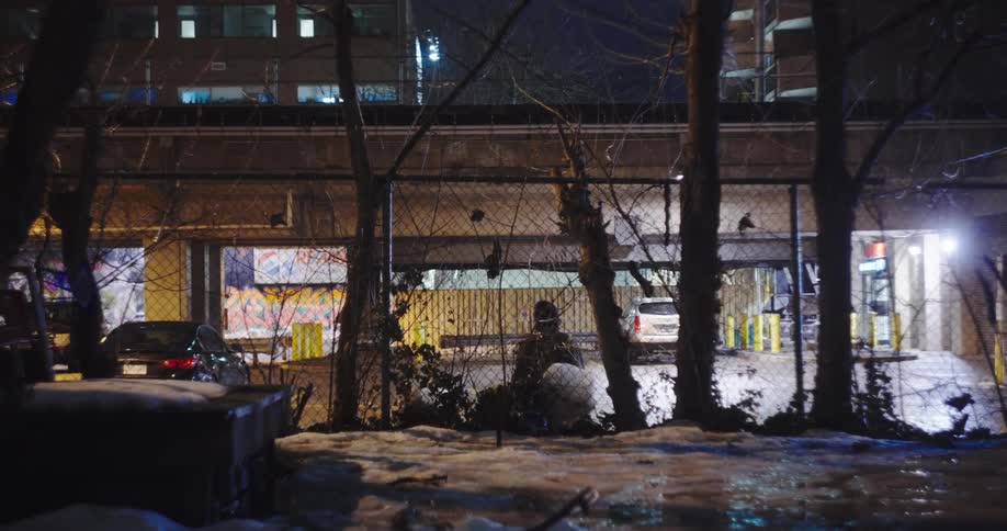 A solitary figure walks along a chain-link fence in a gritty city alley on a cold winter night