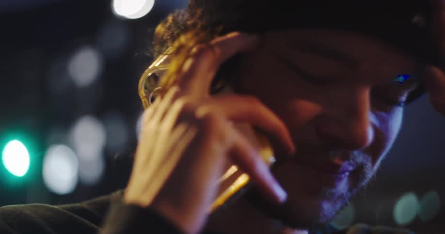 A close-up of a young man in a beanie talking on the phone at night whose happy expression turns to worry upon hearing stressful news