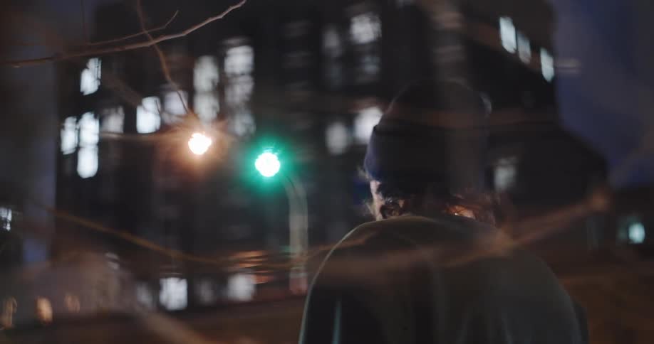 A moody slow-motion shot shows a young man in a beanie walking away down a blurry city street at night