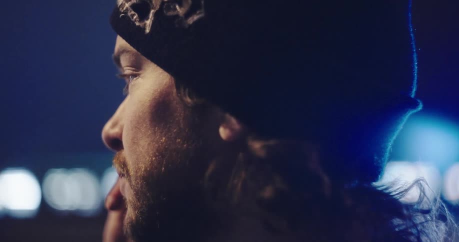 Profile close-up of a serious bearded man in a beanie talking on the phone at night with bokeh city lights in the background