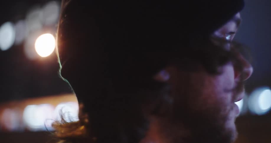 A slow-motion close-up of a thoughtful man in a beanie on a city street at night as the camera pulls focus to a passing train