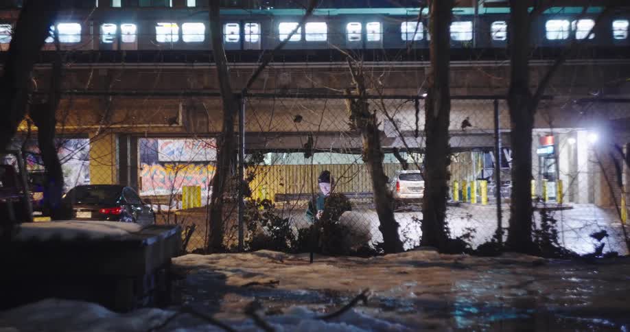 A person sits alone in a snowy, urban landscape at night, illuminated by their phone screen as an elevated train passes in the background