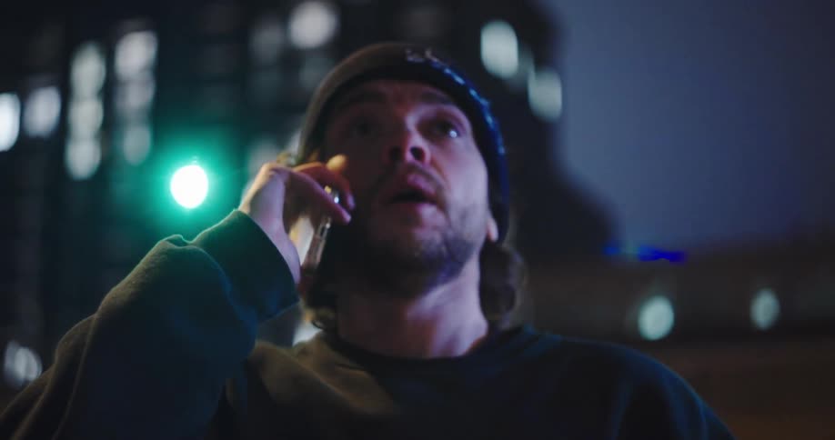A troubled man in a beanie looks stressed while talking on his phone on a city street at night