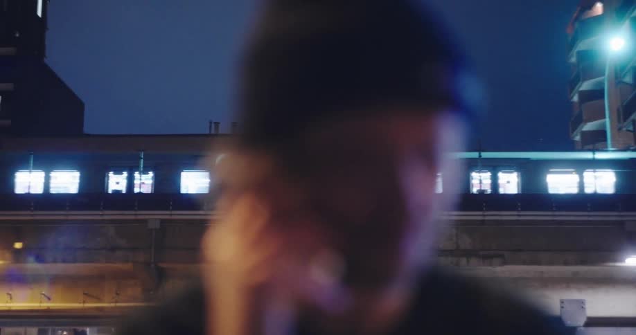 Moody close-up of a young man talking on the phone at night as a brightly lit city train passes in the background