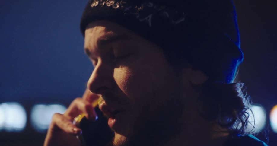 A serious young man in a beanie has an intense phone conversation at night in slow motion, illuminated by blue flashing lights