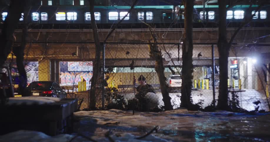 In a gritty city alley on a snowy winter night, a woman is illuminated by her phone while a subway train rumbles by overhead