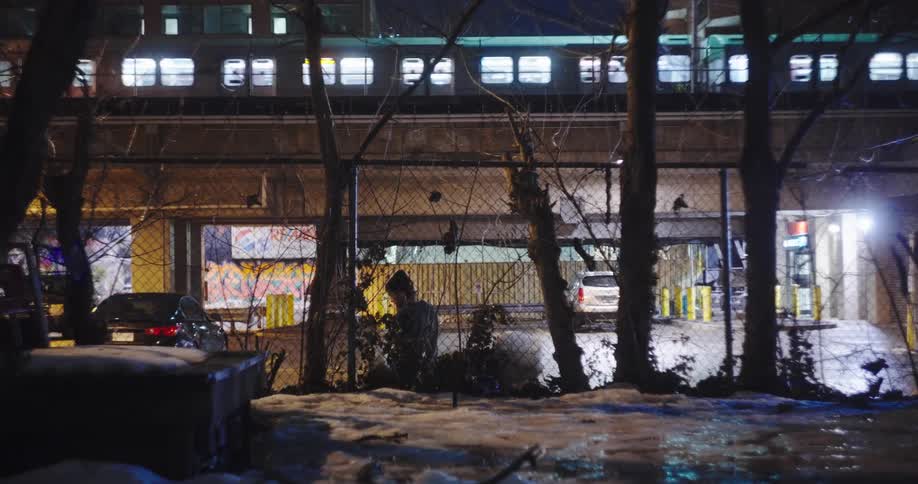 A silhouetted figure watches an elevated train pass by on a cold winter night in a gritty urban landscape