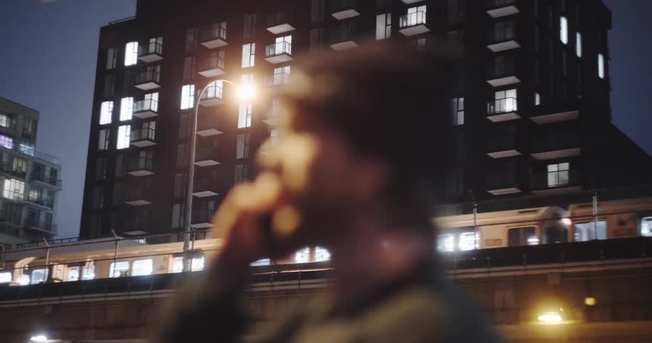 A man talks on his phone as the focus pulls from him to an elevated train passing a modern apartment building at night