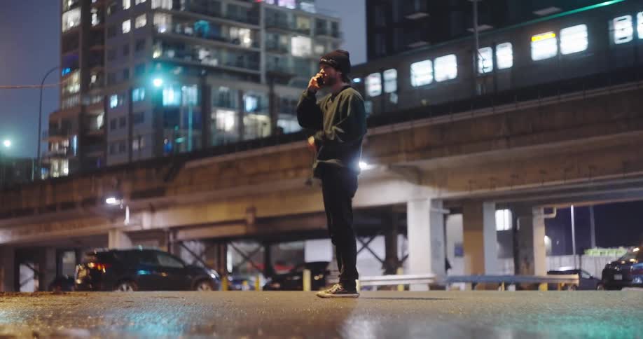 A young man talks on the phone under an elevated train track in the city at night, then hangs up and walks away