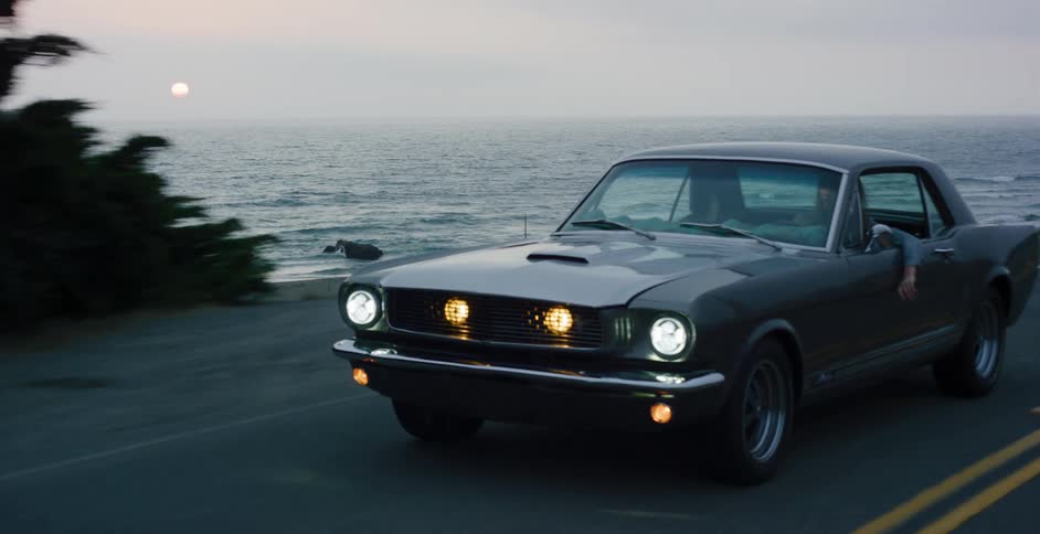 A classic grey Ford Mustang cruises along a scenic coastal highway at dusk with its headlights on