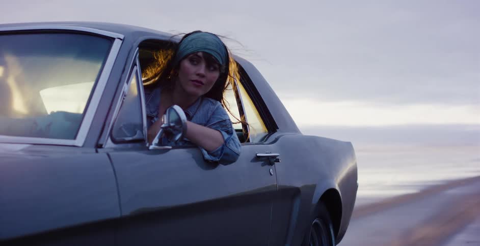 A cool young woman with windblown hair looks back from the driver's seat of a classic car as she drives along the beach at dusk