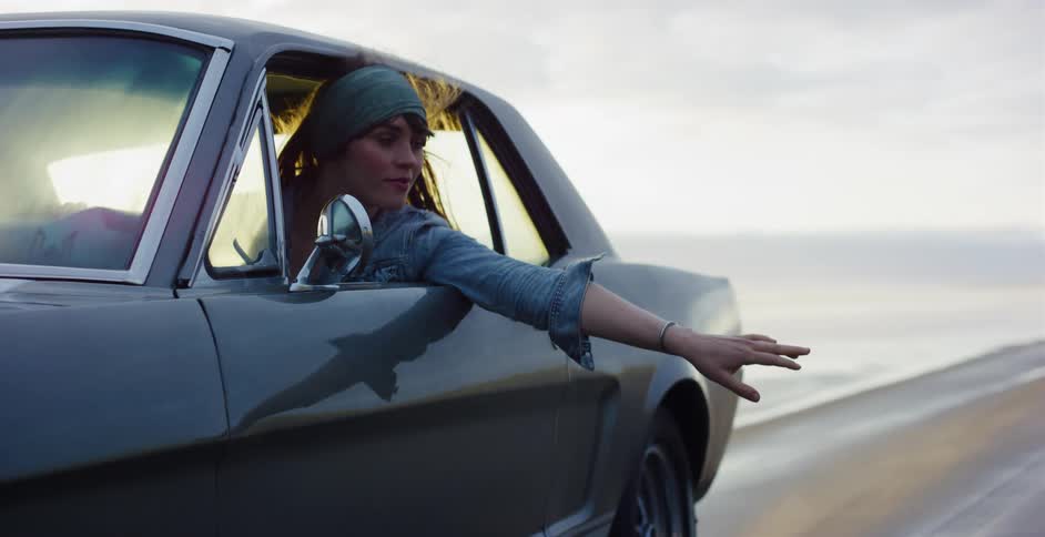 A free-spirited woman in a classic Ford Mustang holds her hand out the window to feel the wind as she drives along the ocean shore