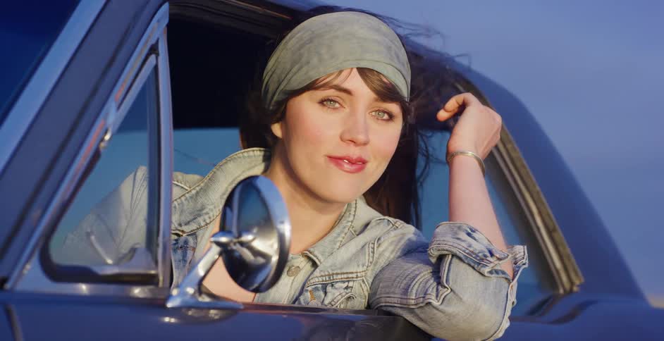 A carefree young woman with a bandana in her hair leans out of a vintage car window, her hair blowing in the wind at golden hour