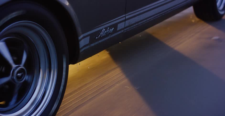 Dramatic close-up of a classic Ford Mustang driving on a wet road at night, with water splashing from the spinning tire