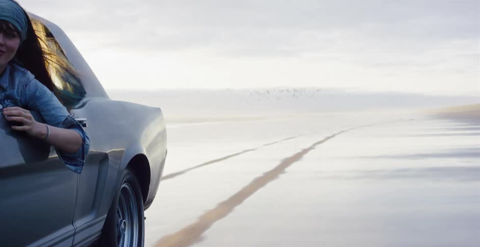 A young woman with wind in her hair enjoys the freedom of driving a vintage muscle car down a wet, sandy beach at sunset