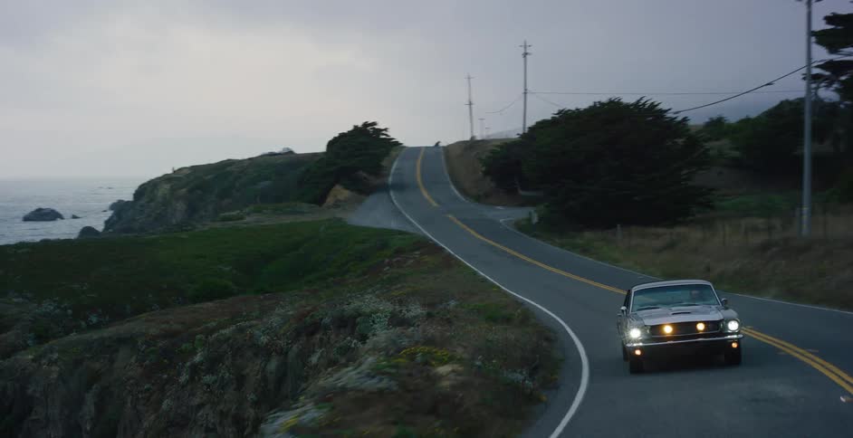 A classic muscle car with its headlights on drives along a winding, scenic coastal highway at dusk