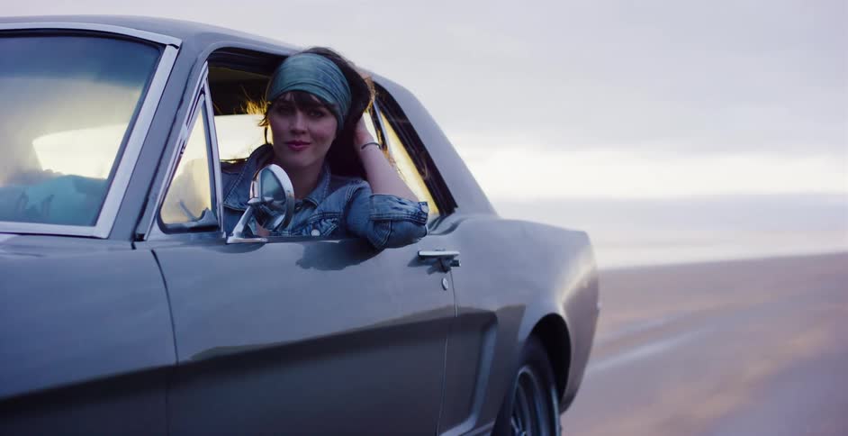 A confident young woman in a classic Ford Mustang looks at the camera as the wind blows through her hair on the beach at sunset