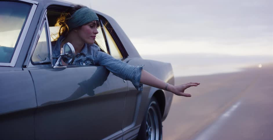 Cinemagraph of a contemplative woman holding her hand out the window of a moving classic car on a beach at dusk