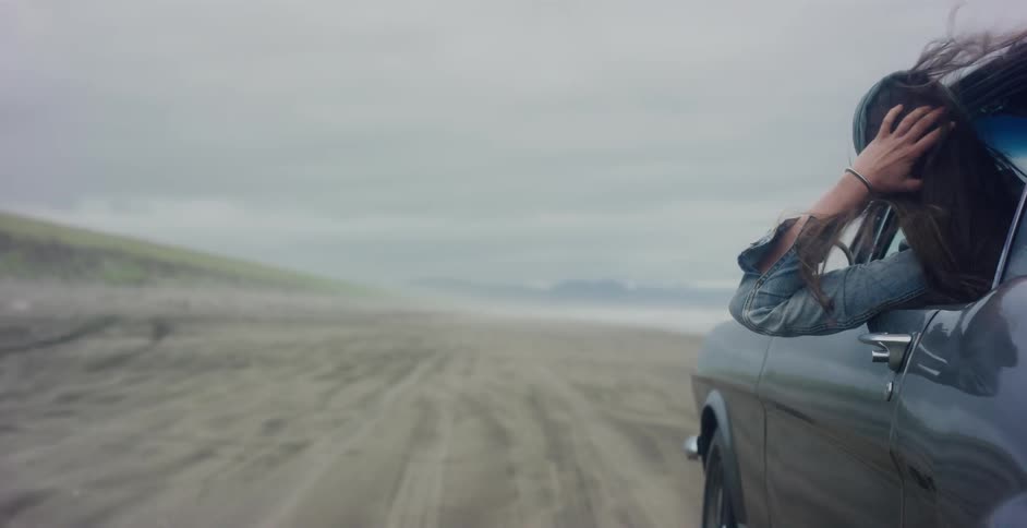 A free-spirited woman with her arm out the window of a vintage car enjoys the wind while driving on a foggy beach in slow motion