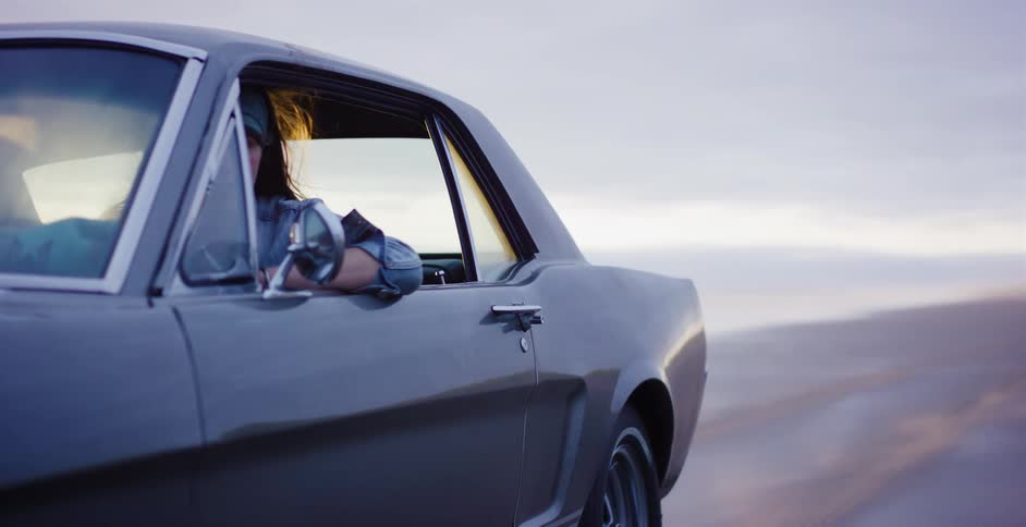 A young woman with wind in her hair drives a classic Mustang across a vast, open salt flat at dusk