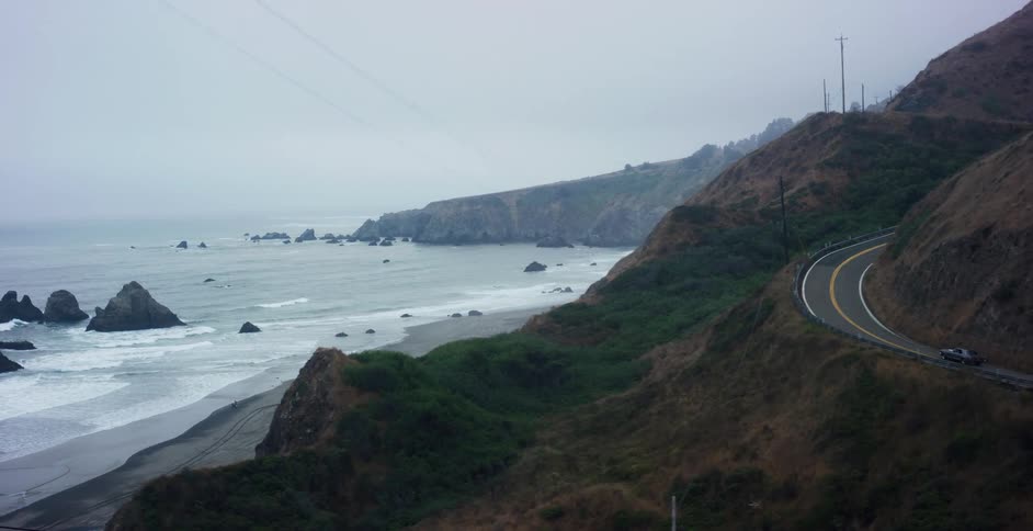 A cinematic tracking shot follows a car on a scenic drive along a winding, foggy coastal highway