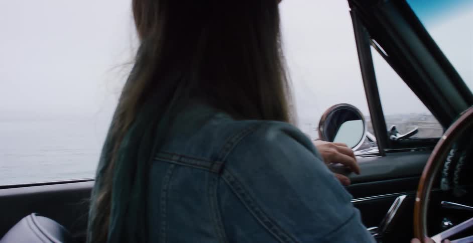 A woman in a classic car drives along the misty coastline in slow motion