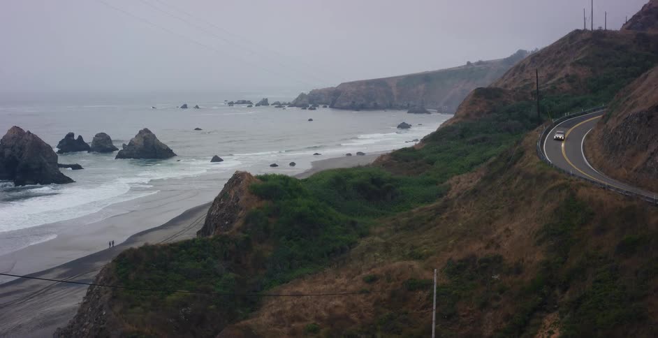 A white car drives on a winding road along a foggy coastline, with waves crashing on the beach below