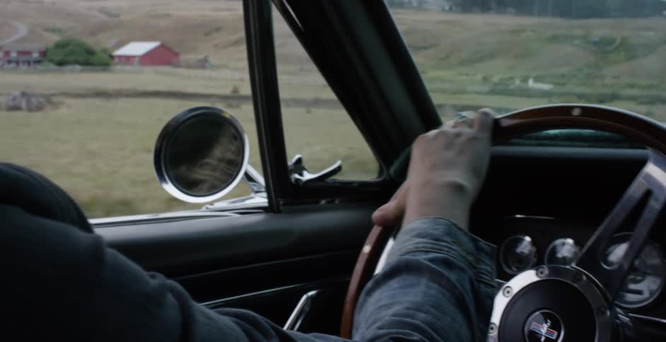 A person's hands grip the wooden steering wheel of a classic car while driving through a scenic rural landscape with a red barn in the distance