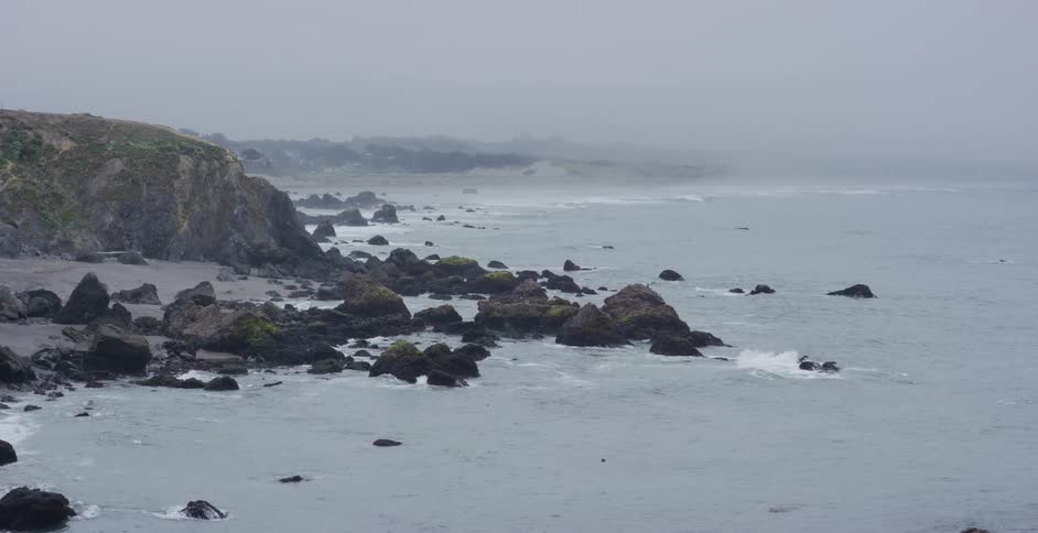 Slow motion waves crash along a rugged, rocky coastline on a very foggy and overcast day