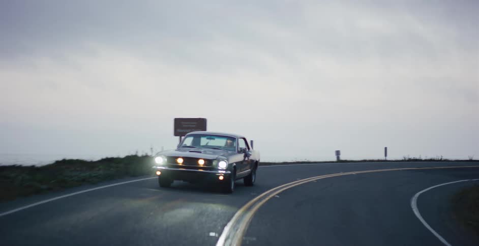 A classic Ford Mustang drives around a bend on a coastal road next to the ocean at twilight