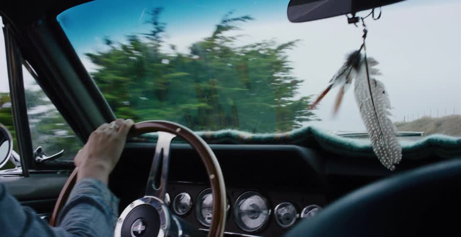 A woman's hands steer a vintage Ford Mustang down a coastal highway as feathers gently sway from the rearview mirror in slow motion