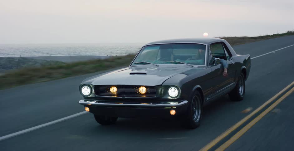 A classic grey Ford Mustang with its headlights on cruises down a scenic coastal highway at dusk