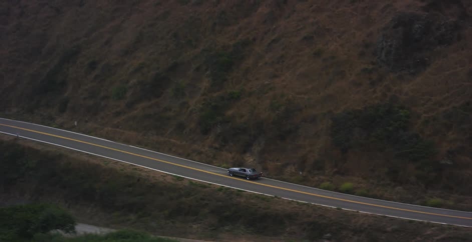 High-angle view of a vintage gray sports car driving along a curving road at the base of a vast, brown mountain