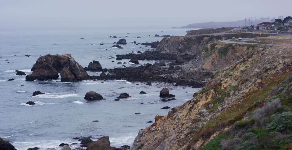A dramatic, slow pan reveals the rocky coastline of the ocean on a gray and overcast day