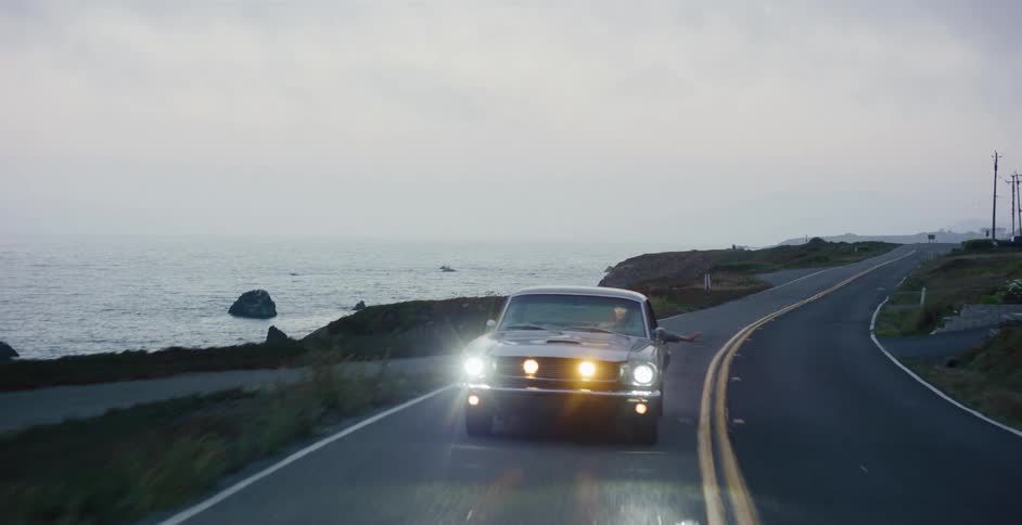 A classic muscle car drives down a winding coastal highway next to the ocean with its headlights on during a gloomy, overcast evening