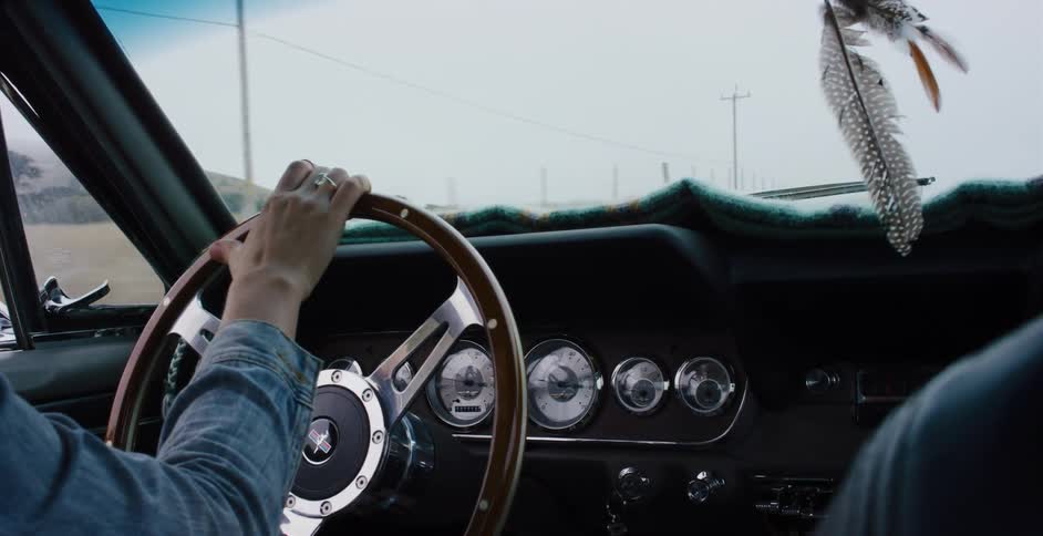 A woman drives a classic car along a foggy coastal road, with feathers swaying from the rearview mirror in slow motion