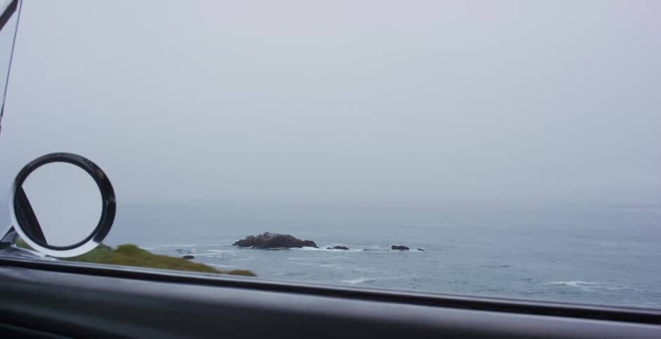 A slow-motion view from a car driving along a foggy coast reveals the vast ocean and a distant rocky island