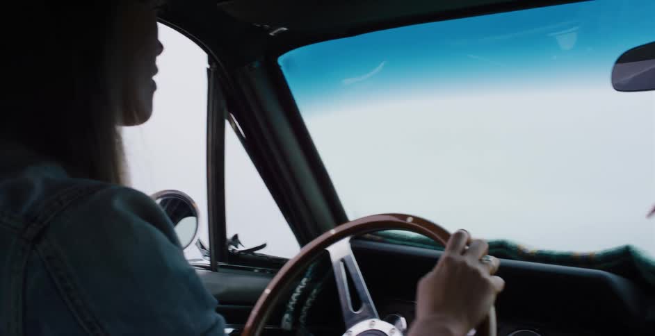 A slow-motion, over-the-shoulder view shows a woman driving a vintage Mustang, with feathers hanging from the mirror and a bright, hazy road ahead