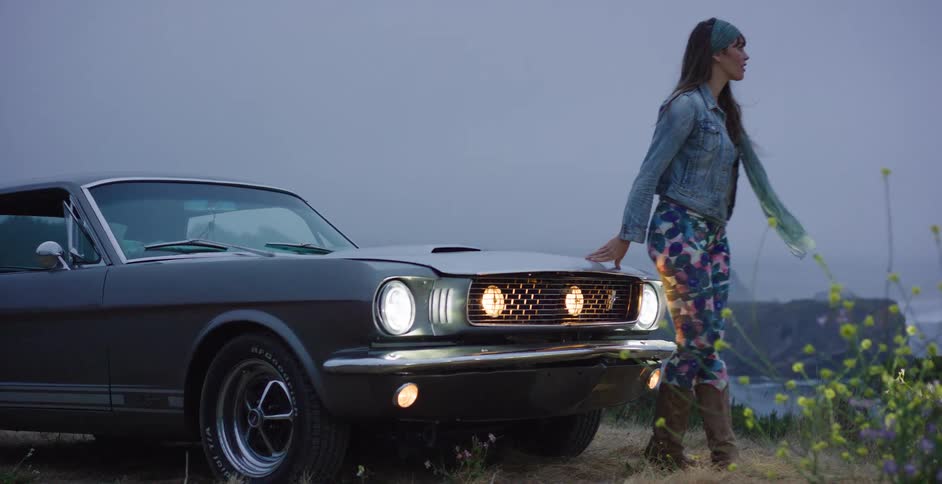 A stylish woman gets out of her vintage Ford Mustang and sits on the hood to admire the foggy coastal view at dusk
