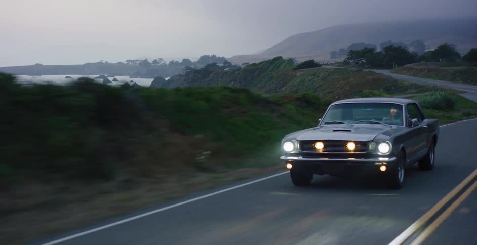 A man drives a classic grey Ford Mustang down a winding coastal highway next to the ocean on a foggy evening