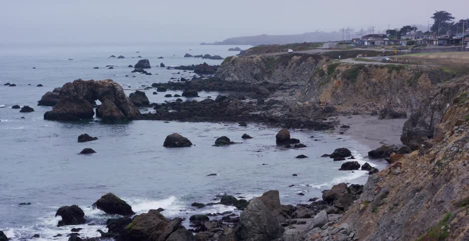A wide, scenic view of a rocky coastline with a natural sea arch on a foggy, overcast day