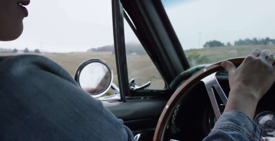 A driver's hands grip the wooden steering wheel of a vintage car during a slow-motion drive on a rural road