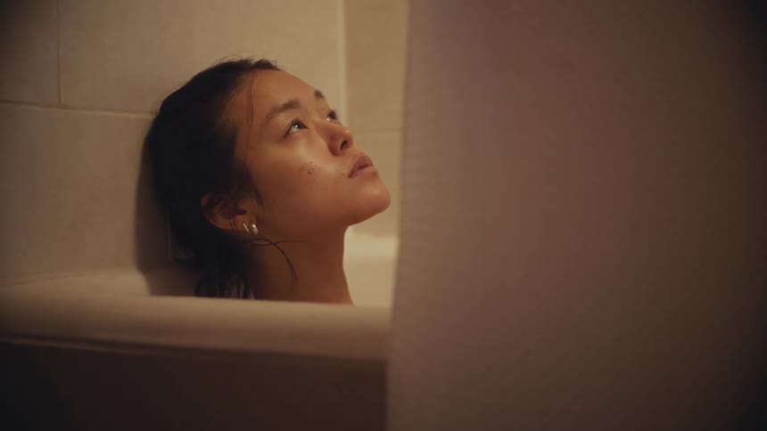 A pensive young woman sits alone in a dimly lit bathtub, looking up thoughtfully