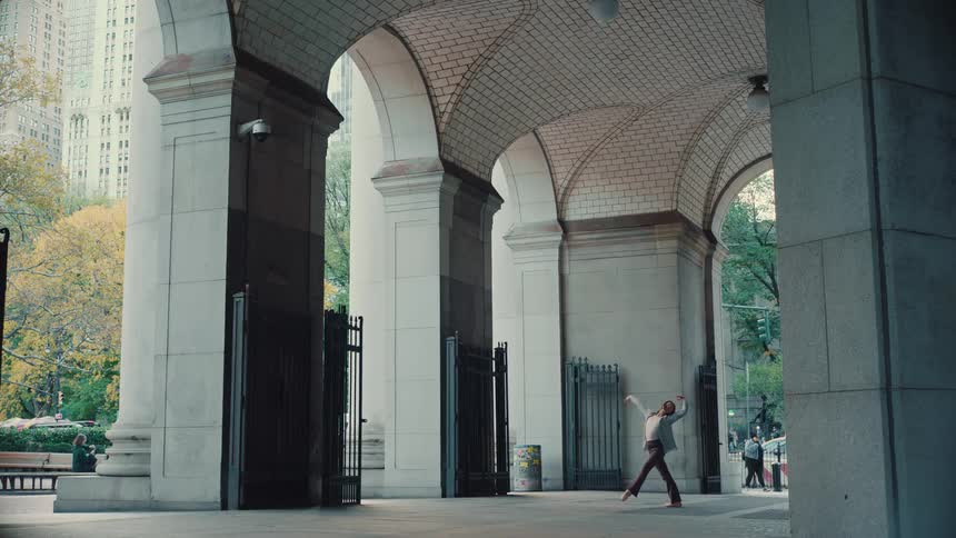 A graceful woman performs a modern dance routine in slow motion under a vaulted stone archway in the city