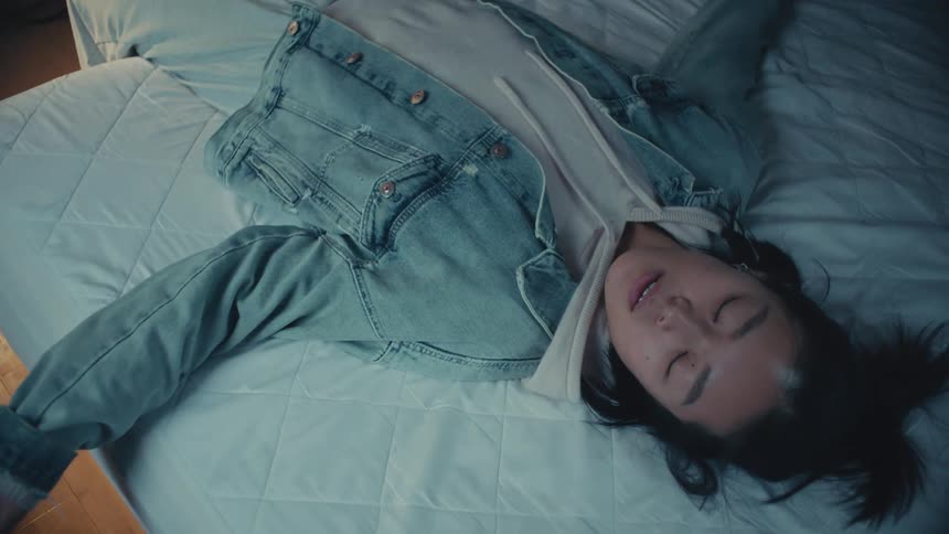 An exhausted young Asian woman in a denim jacket and hoodie falls backward onto her bed in slow motion